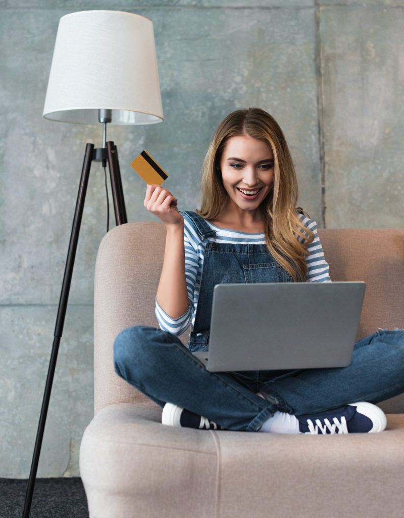 Happy beautiful woman holding credit card in hand and sitting on sofa with laptop.jpg